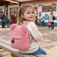 Mädchen mit rosa Teddy-Rucksack mit dem Namen Emma auf einer Bank im Kindergarten