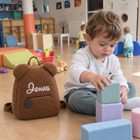 Kleinkind spielt in der Krippe neben seinem braunen Teddy-Rucksack mit dem Namen Jonas