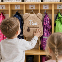 Junge greift nach seinem beigen Teddy-Rucksack mit dem Namen Paul an der Garderobe
