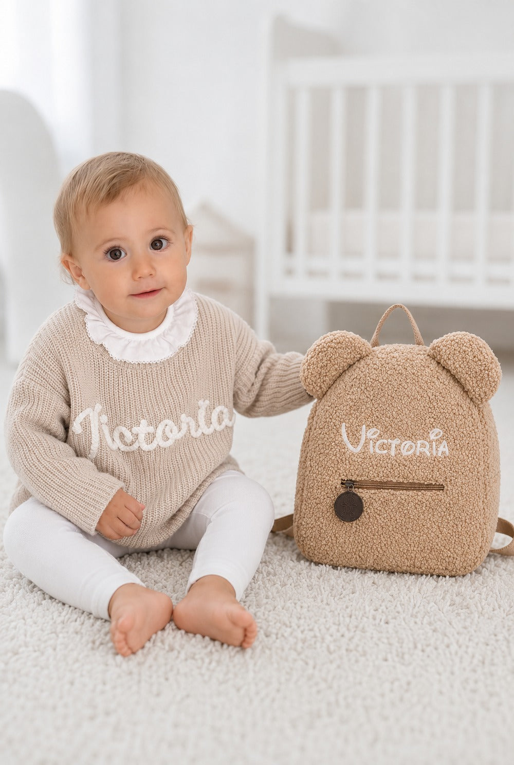 Kleines Mädchen im beigen Babypullover neben ihrem beigen Teddy-Rucksack, beide mit Namen bestickt
