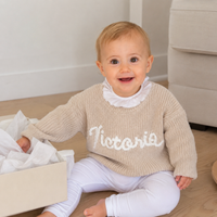 Kleines Mädchen im beigen Babypullover mit handgesticktem Namen Victoria, neben einer geöffneten Geschenkbox