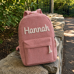 Kundenfoto: rosa Kordsamt-Rucksack mit dem bestickten Namen Hannah, draußen auf einer Steinmauer