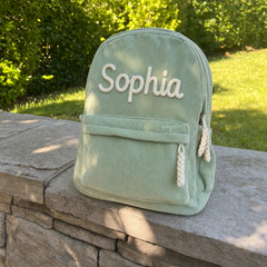 Kundenfoto: salbeigrüner Kordsamt-Rucksack mit dem bestickten Namen Sophia, draußen auf einer Mauer