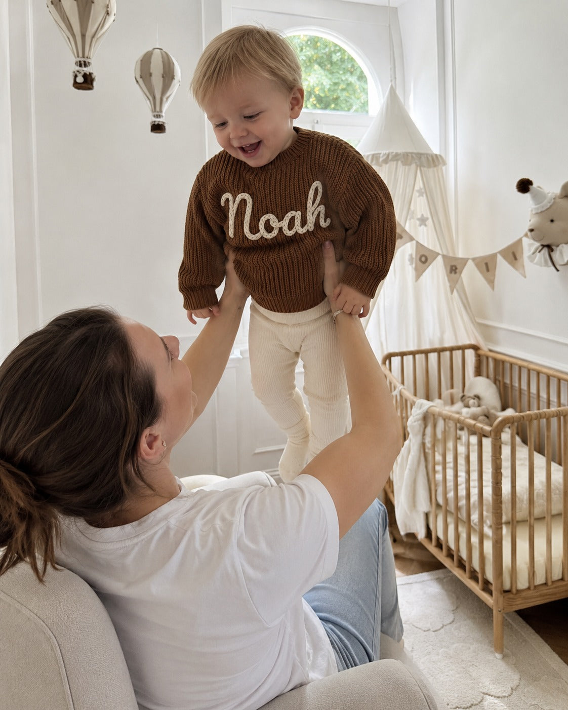 Mutter hebt ihr Kind im Strickpullover mit besticktem Namen Noah hoch