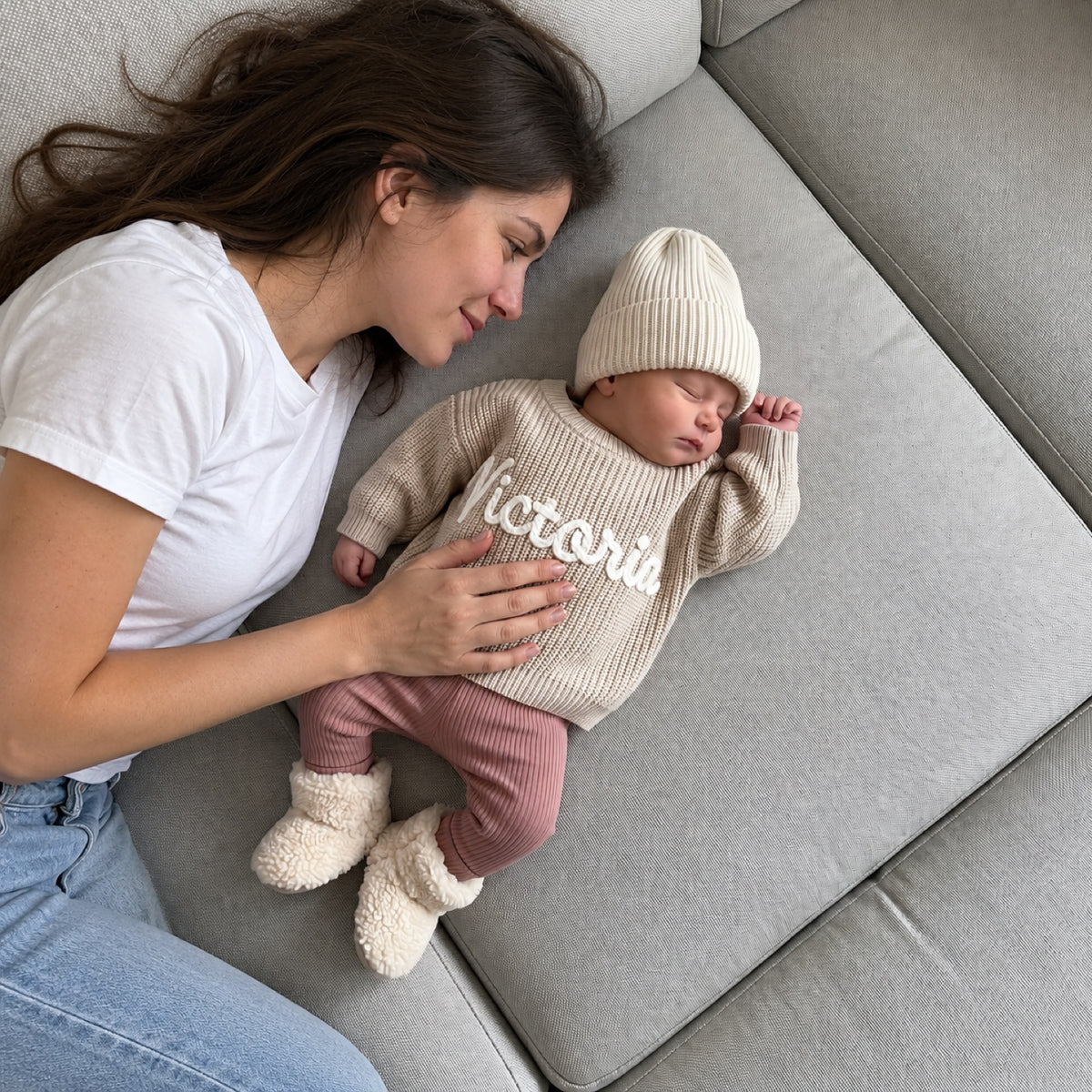 Mutter liegt neben ihrem Neugeborenen im Babypullover mit gesticktem Namen